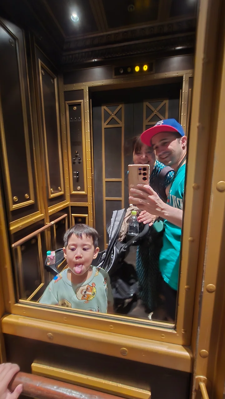Una familia se toma una foto juguetona frente al espejo de un ascensor ornamentado mientras el hijo menor saca la lengua.